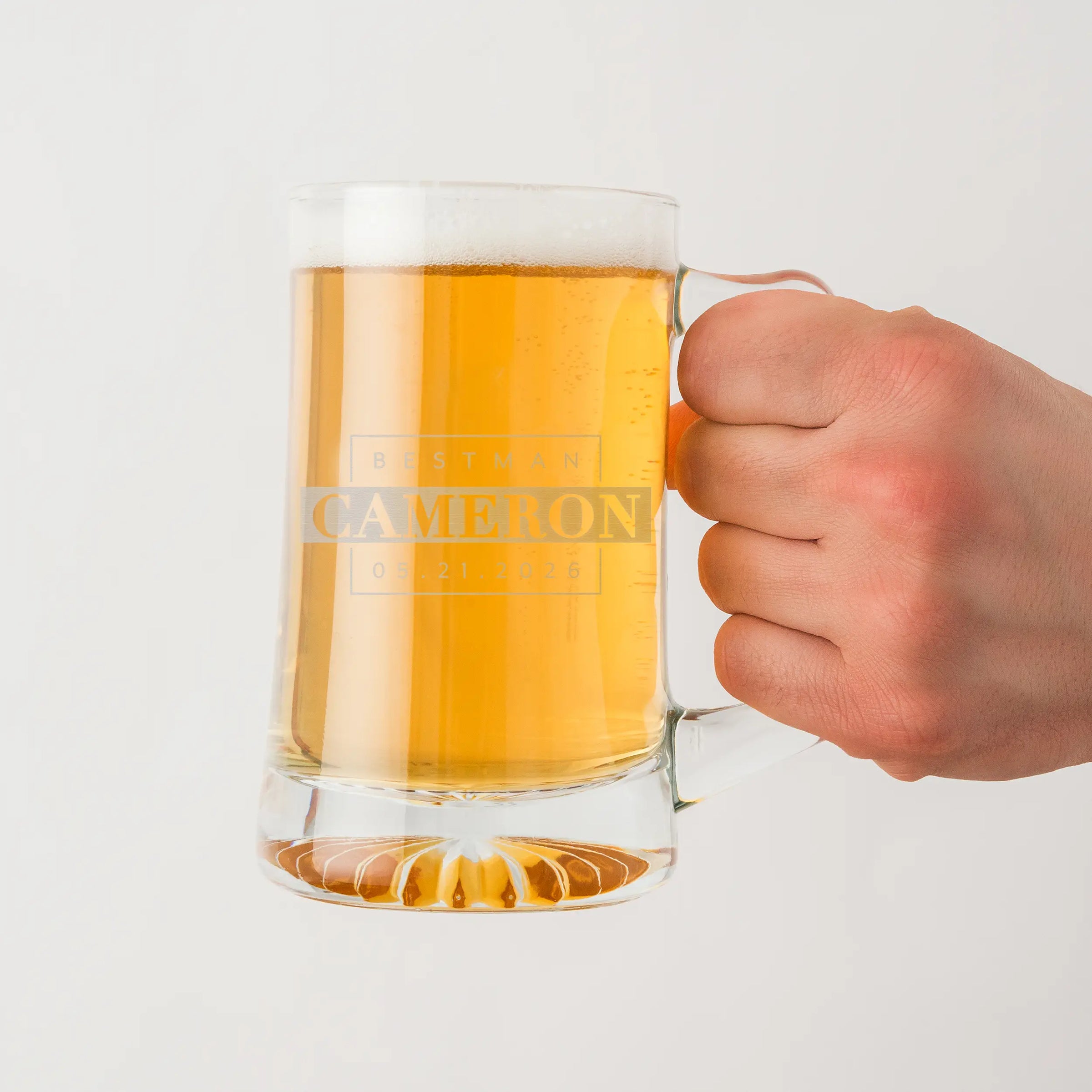Hand holding a glass mug of beer with engraved branding on a light gray background